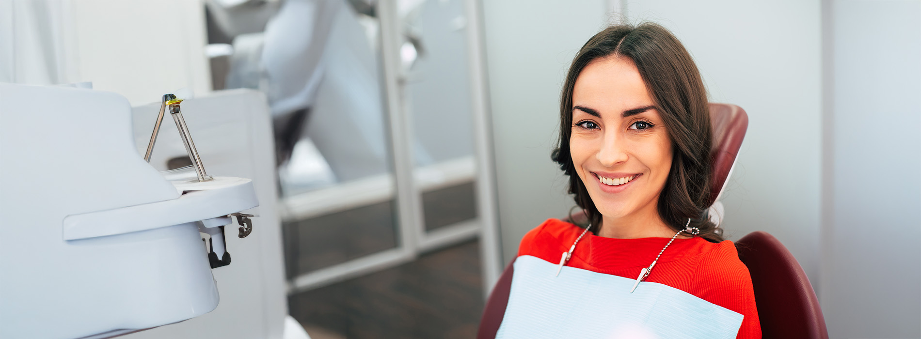 A woman sitting in a dental chair, smiling at the camera, with a dental office setting visible behind her.