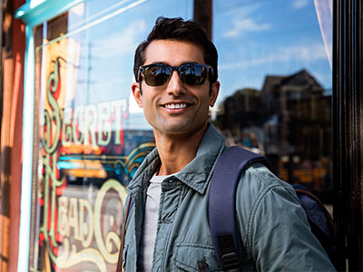 A man with sunglasses and a backpack stands in front of a store window, smiling at the camera.