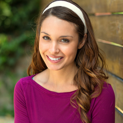 A smiling woman with long hair, wearing a purple top and headband, poses against a wooden fence.