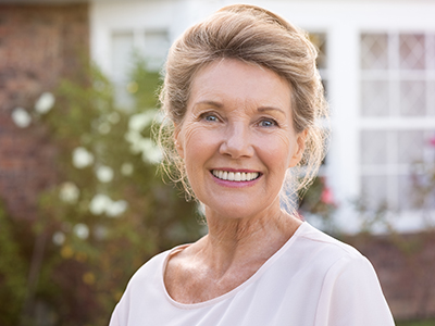 A woman in a white top is smiling and posing outdoors, standing in front of a house with a brick wall and a garden.