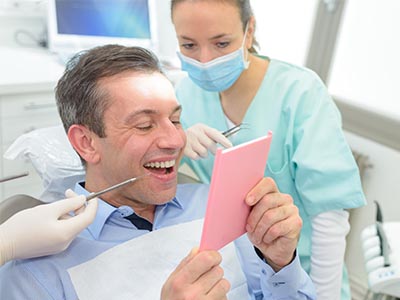 A dentist with a smiling expression, holding up a pink card, surrounded by dental equipment and a woman in scrubs.