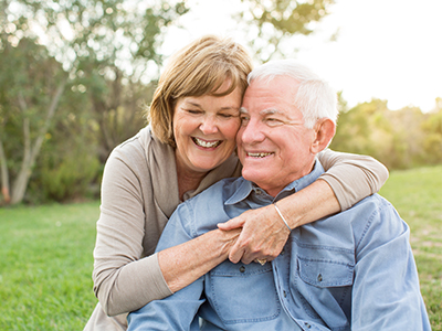 The image depicts an elderly couple embracing each other in a joyful manner, with the man wearing a blue shirt and the woman in a beige top. They are outdoors during what appears to be sunset or early evening, given the soft lighting and shadows.