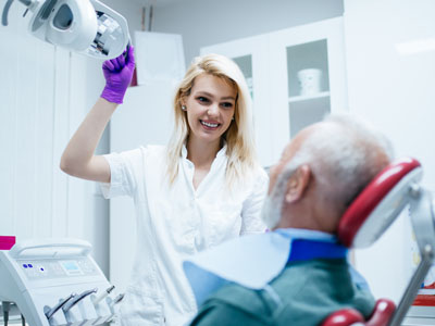 A dental professional, likely a dentist or hygienist, is assisting an elderly person in a dental chair with a smiling expression.