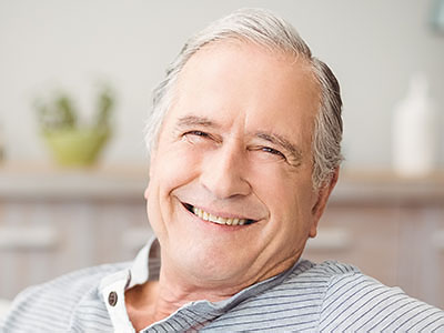 The image shows an older man with gray hair, wearing a dark shirt and light-colored pants, smiling broadly at the camera while seated in a relaxed posture.