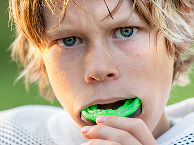 A young boy with blonde hair is seen holding a green toothbrush in his mouth, looking directly at the camera with a neutral expression.